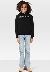 Felpa nera con testo bianco "NEW YORK", abbinata a jeans chiari a zampa d'elefante e scarpe beige. Tessuto testurizzato, vestibilità comoda.