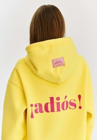 Žltá mikina s kapucňou, ktorá na zadnej strane obsahuje veľký ružový nápis "¡adiós!". Obsahuje aj malú ružovú nálepku na kapucni.