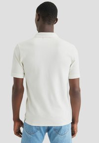 Polo de manga corta blanca, fabricada en un suave tejido texturado, que presenta un cuello clásico y sutiles detalles de costura en la parte posterior.
