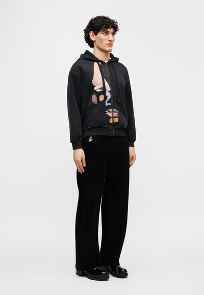 Μαύρο φούτερ με κουκούλα και αφηρημένα γραφικά σχέδια σε παστέλ χρώματα, κλείσιμο με φερμουάρ και ριμπ τελείωμα, σε συνδυασμό με μαύρο παντελόνι από κοτλέ ύφασμα και παπούτσια.