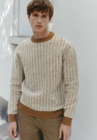 Textured cream sweater with a honeycomb pattern, featuring a brown ribbed collar and cuffs. Paired with beige trousers.