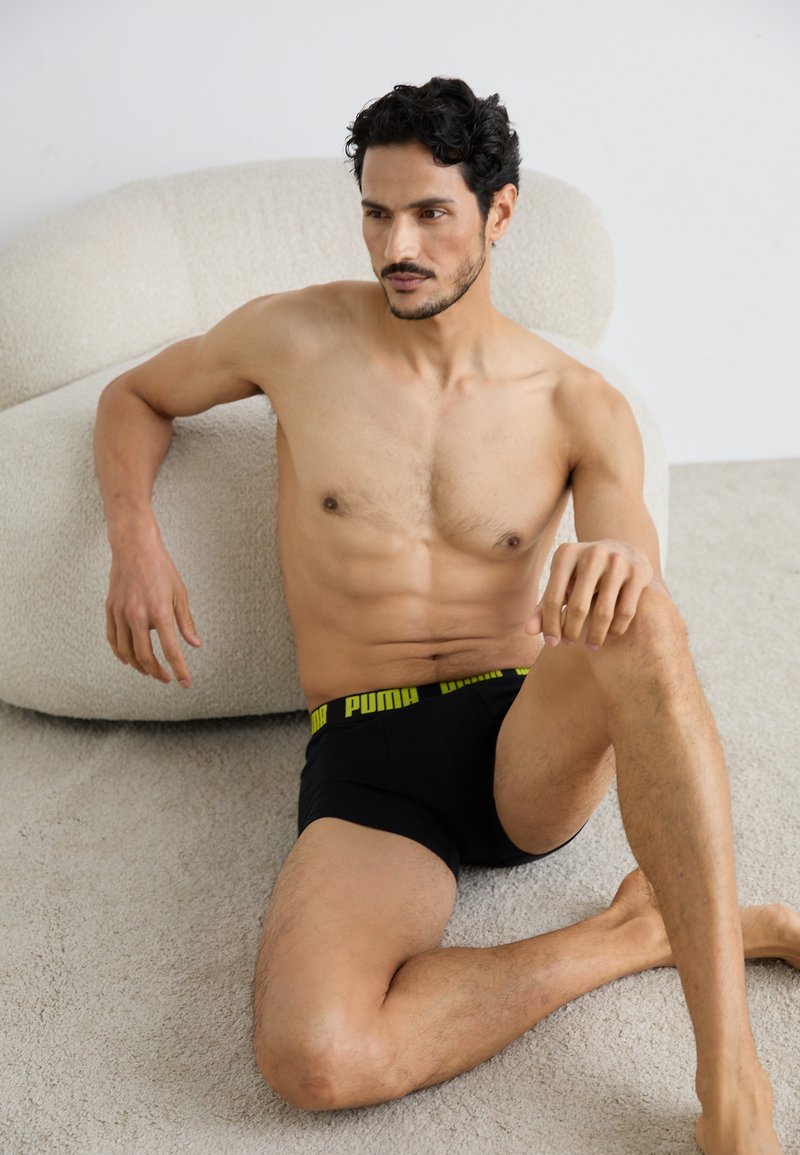 Boxeri negri cu un brâu cu logo Puma galben, purtați de un bărbat fără cămașă, așezat pe un covor moale de culoare deschisă, alături de un scaun moale, rotunjit.