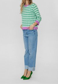 Pull à motifs verts et blancs avec bordure violette, associé à un jean bleu clair à coupe décontractée et chaussures à talons pointus verts.