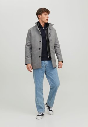 Abrigo de mezcla de lana gris con cremallera frontal, grandes botones y cuello alto. Lleva puesto sobre un suéter azul marino y combinado con jeans claros y zapatillas.