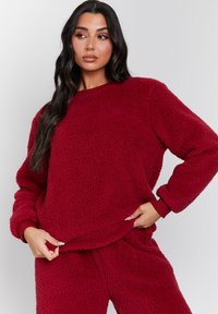 Kvinna med långt mörkt hår som bär en matchande röd sherpa fleece-tröja och byxor, håller ner tröjans fåll mot en enkel ljus bakgrund.