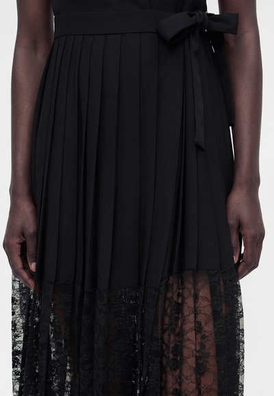 Μαύρη πλισέ φούστα με δαντελένιο τελείωμα και δεμένη μέση, φορεμένη από άτομο με σκουρόχρωμο δέρμα, εμφανιζόμενη από τη μέση έως τα γόνατα.