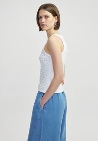 Weißes, ärmelloses Oberteil mit strukturiertem Design und kleinen Perforationen. Kombiniert mit hellblauen, locker sitzenden Jeanshosen mit Taschen.