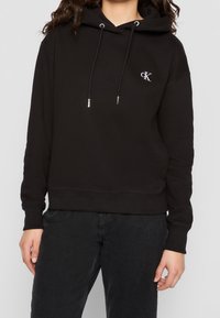 Personne portant un sweat à capuche noir avec cordons de serrage et petit logo blanc "CK" sur la poitrine, associé à un pantalon noir.