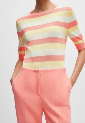 Kobieta w pastelowym, paskowanym dzianinowym topie z krótkimi rękawami, włożonym do wysokich, łososioworóżowych spodni z rękami w kieszeniach.