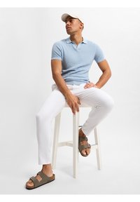 Himmelblaues, strukturiertes Poloshirt, weiße Slim-Fit-Hosen und braune Sandalen mit zwei verstellbaren Riemen, auf einem weißen Hocker vor einem schlichten Hintergrund sitzend.