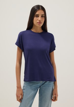Giovane donna con lunghi capelli scuri che indossa una t-shirt blu navy a maniche corte e jeans azzurri, in piedi davanti a uno sfondo semplice.
