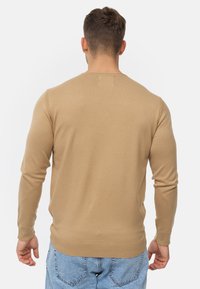 Beige strikket sweater med lange ærmer og rund hals. Har ribbede kanter og manchetter. Glat tekstur med minimal sømdetaljering på bagsiden.