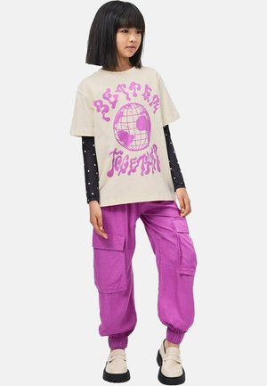 T-shirt graphique beige avec inscription rose « Better Together », associé à un pantalon cargo rose avec poches latérales et poignets élastiques, porté avec des chaussures plates camel.