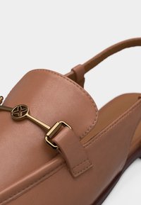 Καφέ δερμάτινο μοκασίνι με χρυσή μεταλλική λεπτομέρεια σε σχήμα αλόγου και λουράκι τύπου slingback σε ουδέτερο φόντο.