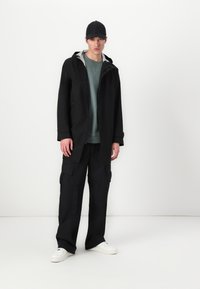 Zwarte cargojack met capuchon, laag over een groene sweatshirt, aangevuld met zwarte cargobroeken en witte sneakers. Vlakke pet als accessoire.