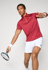 Red athletic polo shirt with striped texture, yellow collar accents, and logo; paired with white shorts and holding a tennis racket.