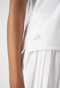 Bílý sporty top s volným střihem, zdobený jemným šedým logem Adidas a bočními rozparky pro snadnější pohyb, vyrobený z lehké tkaniny.