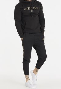 Hombre vistiendo un hoodie negro de SikSilk y joggers a juego con letras doradas, zapatillas blancas, manos en los bolsillos, de pie contra un fondo blanco.