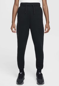 Pantalon de survêtement noir en tissu doux, avec taille à cordon, poches latérales zippées, jambes effilées et poignets côtelés.
