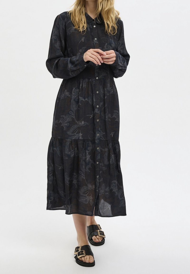 Kvinna klädd i en mörk, knapprad skjortklänning i midi-längd med subtilt blommönster och svarta sandaler med guldspännen, som står framför en vit bakgrund.