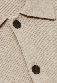 Beige ribbad polotröja med krage, som har mörkbruna knappar och en mjuk textur.