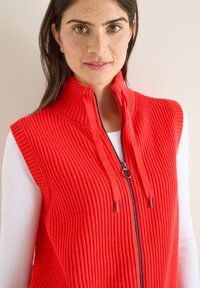 Rotes, geripptes, ärmelloses Weste mit hohem Kragen, Frontreißverschluss und Kordelzug. Über einem weißen Langarmshirt getragen, was die Struktur hervorhebt.