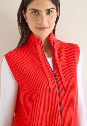 Rotes, geripptes, ärmelloses Weste mit hohem Kragen, Frontreißverschluss und Kordelzug. Über einem weißen Langarmshirt getragen, was die Struktur hervorhebt.