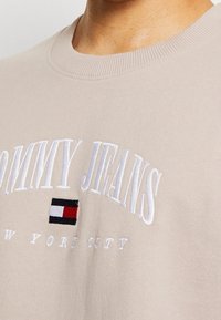 Beżowa bluza z haftowanym napisem „TOMMY JEANS” oraz małą flagą w kolorach czerwonym, białym i niebieskim. Prążkowany, okrągły dekolt i miękka w dotyku tkanina.