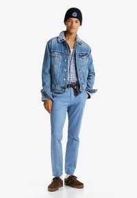 Veste en denim avec de grandes poches, chemise à carreaux bleue, pantalon bleu clair et chaussures marron. Bonnet noir avec un détail de logo.