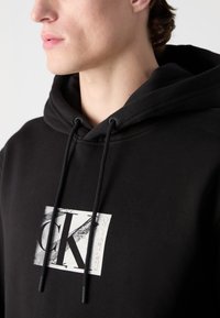 Black hoodie featuring a central white graphic with "CK" and text "Calvin Klein Jeans." It has a drawstring hood and soft fabric texture.