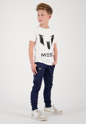 T-shirt blanc avec logo noir et accents, associé à un pantalon de jogging bleu marine avec poches latérales et logo discret. Porté avec des baskets blanches.