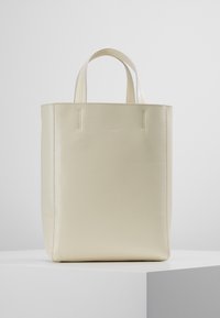 Krämfärgad läder toteväska med strukturerad design, platt botten och dubbla korta handtag. Slät textur med minimala sömnadsdetaljer.