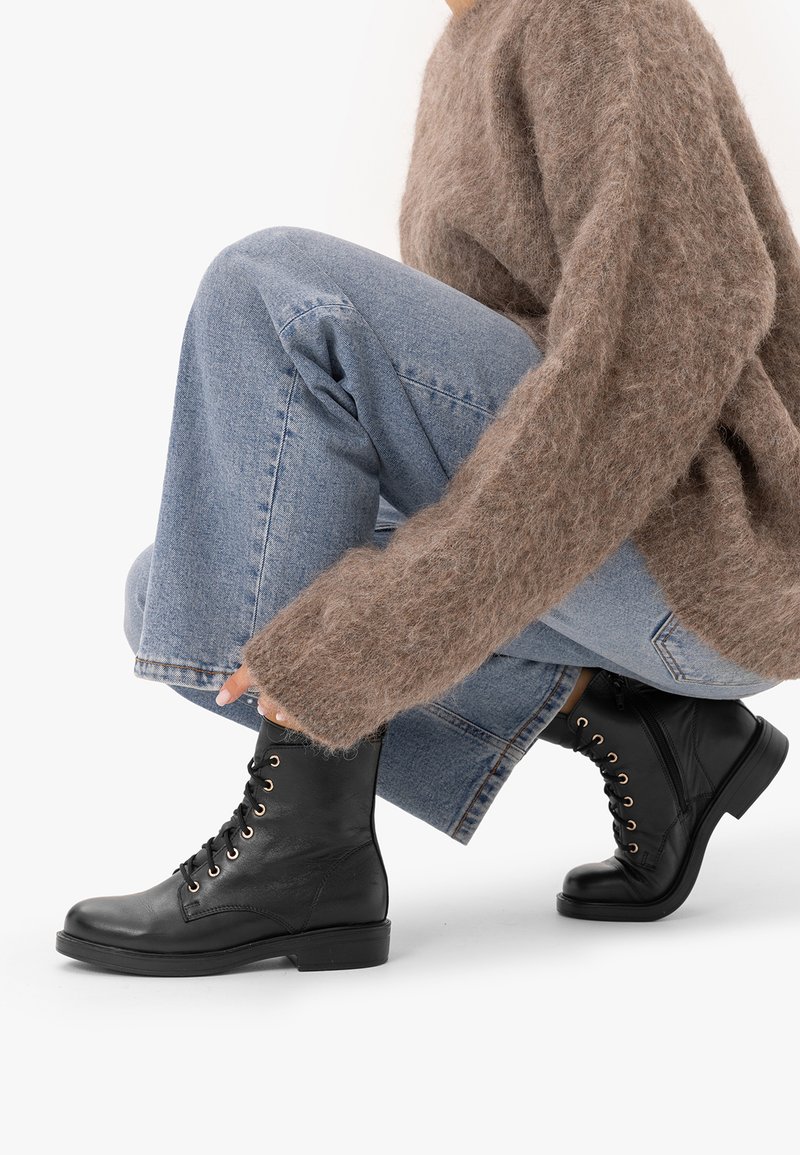 Personne portant un jean bleu clair, un pull marron duveteux et des bottines noires à lacets, accroupie avec une main près de la chaussure.