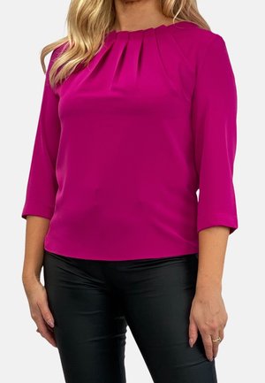 Nainen pukeutuneena fuksianpunaiseen puseroon, jossa on laskoksia pääntiellä ja kolme neljänneksen hihaa, yhdistettynä mustiin istuviin housuihin.