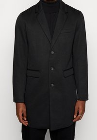 Cappotto di lana nera con un design su misura, caratterizzato da una chiusura a un solo petto, due tasche frontali e un rever a punte. Tessuto liscio, lunghezza al ginocchio.