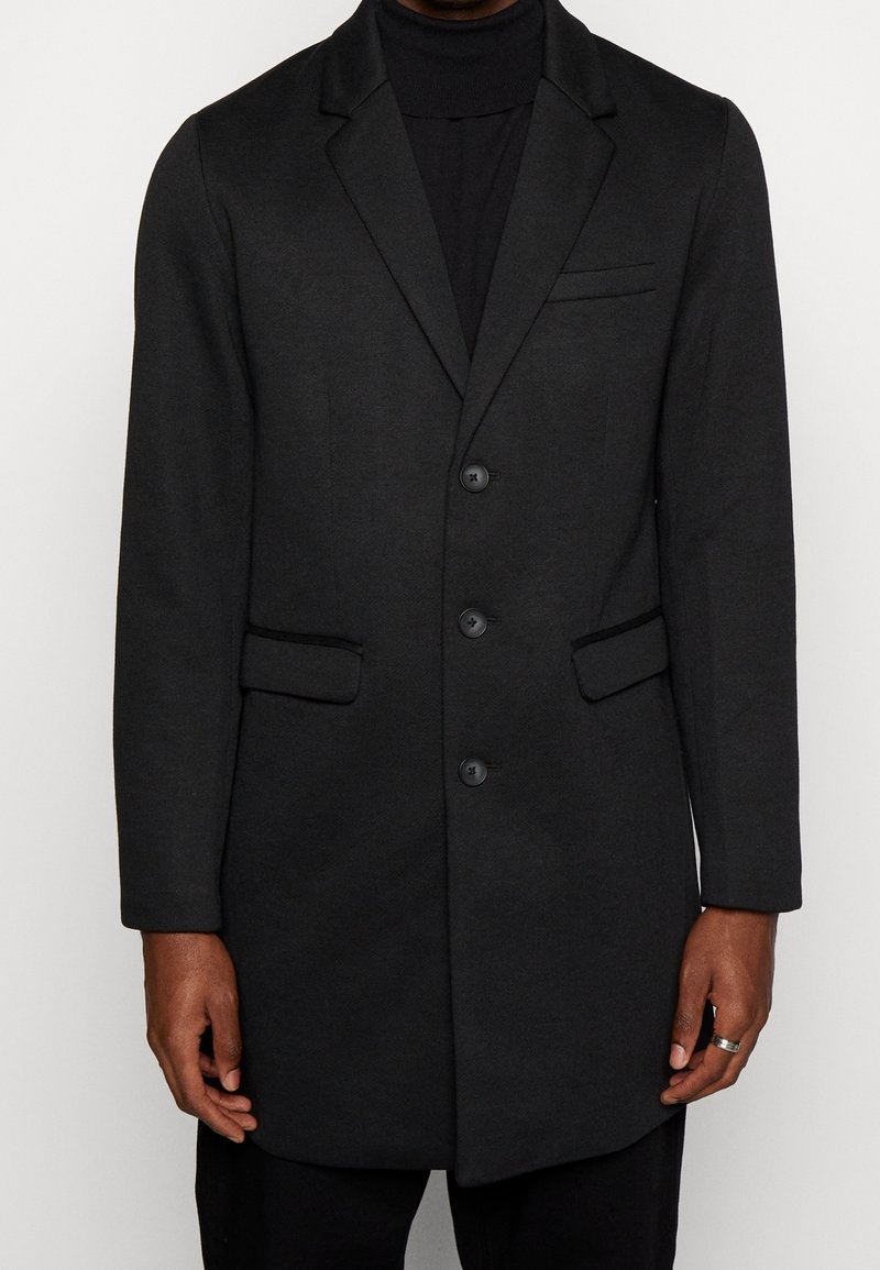 Cappotto di lana nera con un design su misura, caratterizzato da una chiusura a un solo petto, due tasche frontali e un rever a punte. Tessuto liscio, lunghezza al ginocchio.