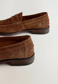 Mocasines de ante marrón con una textura suave, costuras ligeramente elevadas y un sólido tacón de bloque de madera oscura. Diseño clásico tipo slip-on.