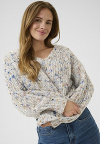 Jonge vrouw met lang bruin haar, gekleed in een gekleurde chunky gebreide trui en blauwe spijkerbroek, glimlachend met haar handen in de trui gestoken.