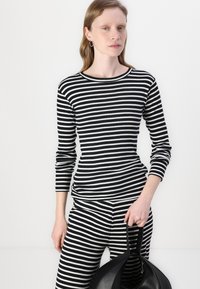 Schwarz-weiß gestreiftes Langarmoberteil und passende Hose, aus einem gerippten Stoff. Das Model hält eine schwarze Handtasche mit geschwungenen Griffen.