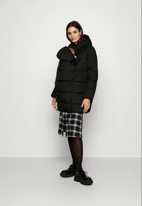 Žena u crnom puffer jakni s ugrađenim nosiljkom za bebe, drži dojenče, uparenom s kariranom suknjom, crnim tajicama i platformama.
