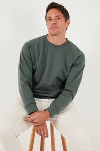 Felpa verde realizzata in un materiale morbido, con scollatura rotonda, maniche raglan e un piccolo dettaglio con logo. Abbinata a pantaloni chiari.