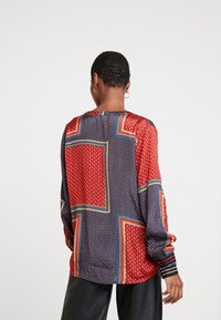 Blouse à manches longues en rouge, marine et crème avec des motifs géométriques. Tissu lisse avec une coupe décontractée et un ourlet arrondi.