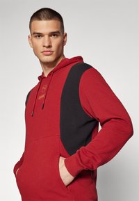 Felpa rossa con pannelli laterali neri, cappuccio con cordino e tasca a marsupio. Tessuto liscio e design casual, con un piccolo logo sul davanti.