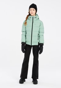 Helles grünes Puffer-Jackett mit Kapuze, schwarzen Reißverschlüssen, kombiniert mit schwarzen gemusterten Hosen und Handschuhen, getragen mit einer schwarzen Wollmütze.