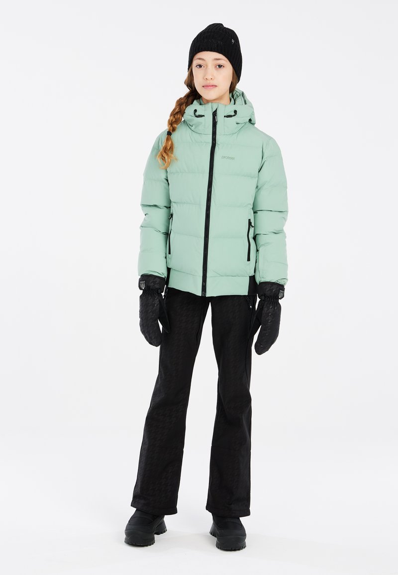 Helles grünes Puffer-Jackett mit Kapuze, schwarzen Reißverschlüssen, kombiniert mit schwarzen gemusterten Hosen und Handschuhen, getragen mit einer schwarzen Wollmütze.