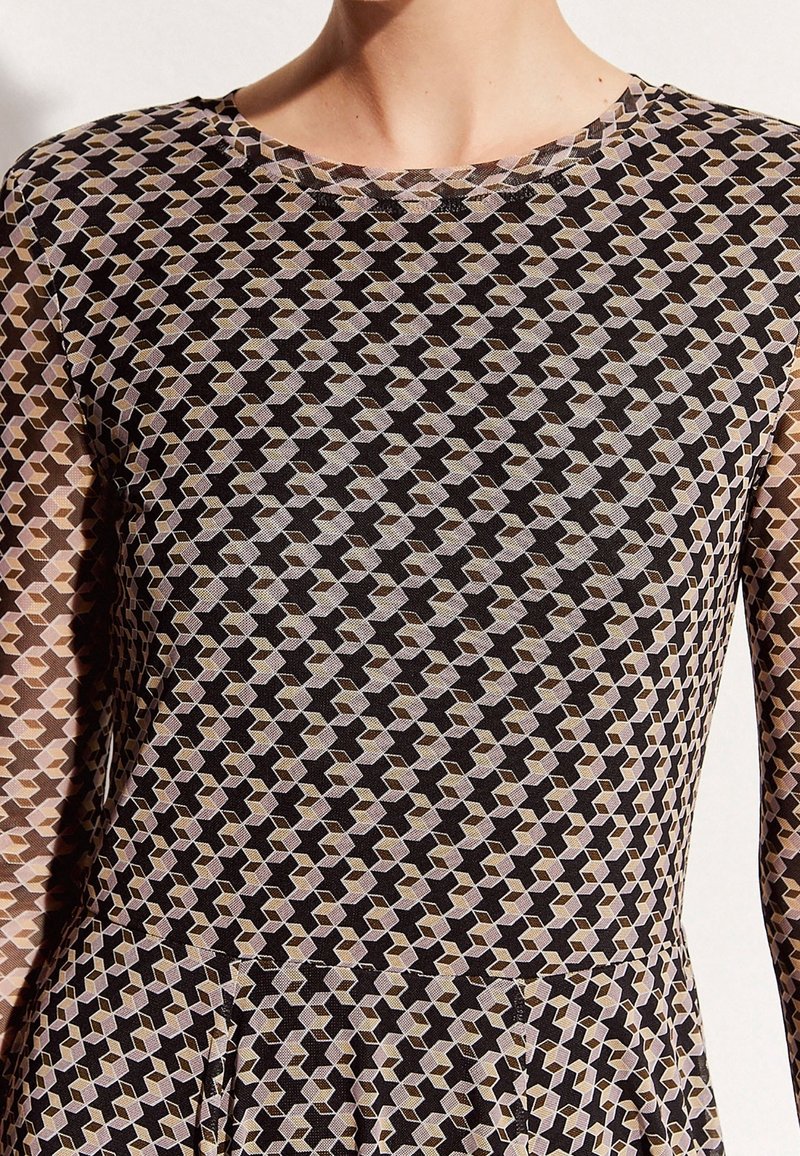Femme portant une robe à manches longues avec un motif géométrique de formes emboîtées noires, beiges et violet clair.