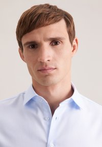 Camicia azzurro chiaro con tessuto a trama strutturata, caratterizzata da colletto appuntito e pattina con bottoni visibili, indossata da un modello maschile su sfondo neutro.