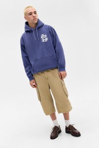 Lila Hoodie mit weißem Grafikdesign, Fronttasche und Kordelzug. Beige Cargo-Shorts mit Seitentaschen. Braune Schnürschuhe.
