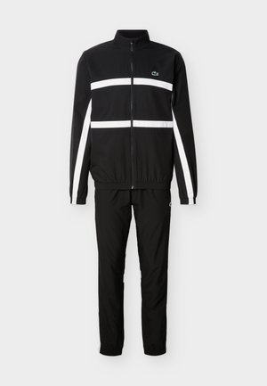 Survêtement noir avec veste à fermeture éclair intégrale présentant des rayures blanches horizontales et verticales, et pantalon noir assorti avec poignets élastiques.