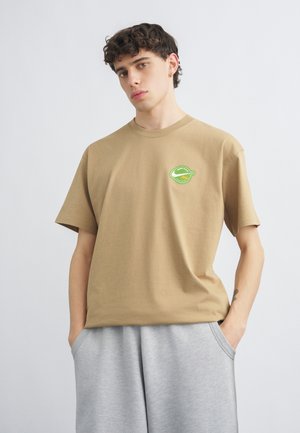 Noor mees lokkis juustega, kannab beežikat T-särki rohelise logoga, halli lühikesi pükse, üks käsi taskus, seisab valge tausta ees.